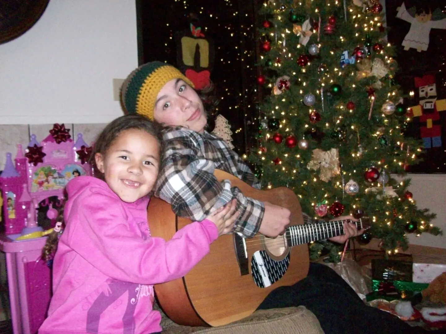 A girl and boy playing guitar in front of christmas tree.